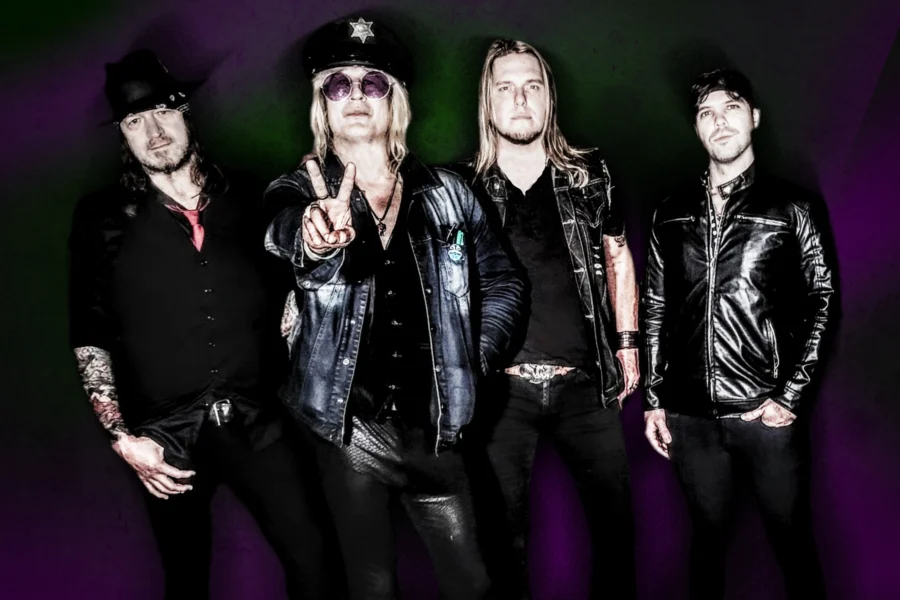 Image of the rock band Enuff Z' Nuff standing together with dark purple and green background. Lead singer Chip Z'Nuff with hand up in a peace sign.